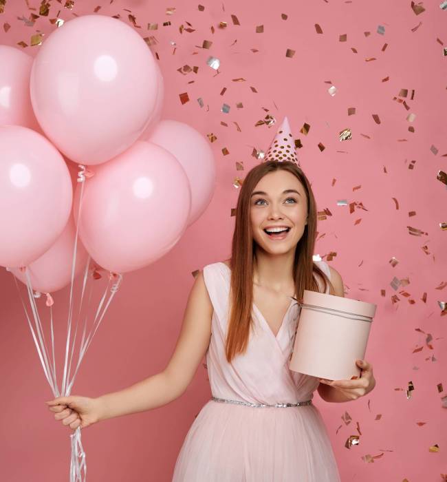 Beautiful smiling woman in dress and birthday hat holding gift box and pastel pink air balloons and falling confetti against pink background. cute happy young girl celebrating birthday party.